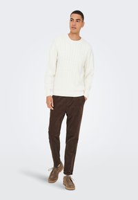 Weißer Pullover mit Zopfmuster, braune Cordhosen und braune Schuhe. Der Pullover hat gerippte Bündchen und Saum. Die Hose ist im geraden Bein geschnitten.