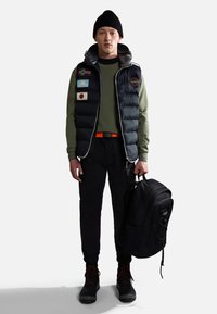 Dunkelblaues Pufferwesten mit Aufnähern, grüner Pullover, schwarze Jogginghose, orangefarbener Gürtel und robuster schwarzer Rucksack. Hat schnürbare Stiefel.