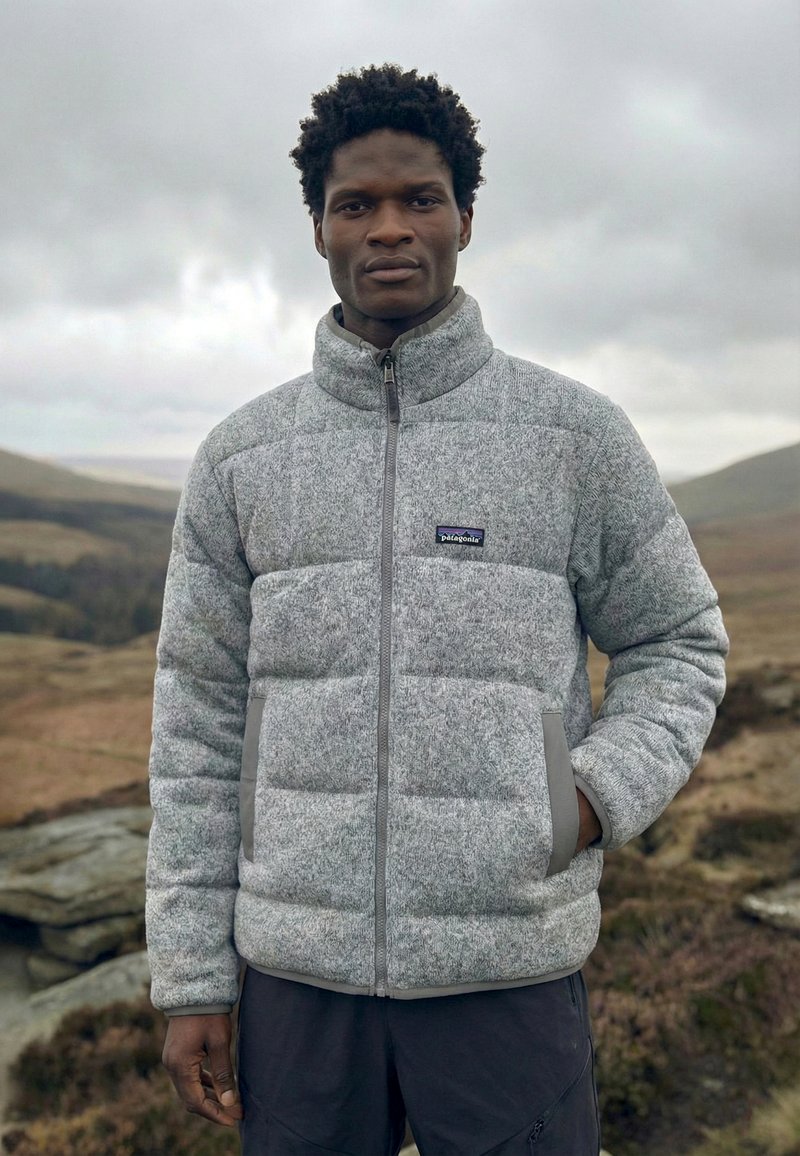 Muž stojící venku v šedé péřové bundě Patagonia, s rukama v kapsách, v pozadí zatažená obloha a zvlněné kopce.