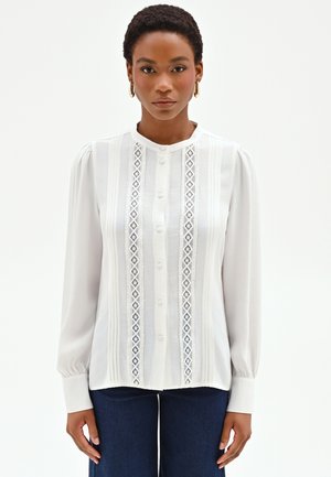 Donna che indossa una camicetta bianca con pannelli in pizzo e maniche lunghe, abbinata a pantaloni blu scuro, in piedi davanti a uno sfondo semplice.