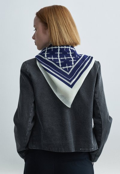 Τετράγωνο φουλάρι σε ναυτικό μπλε και λευκό, με γεωμετρικά σχέδια και τριγωνικό μοτίβο, φορεμένο πάνω από ένα σκούρο γκρι σακάκι με υφή.