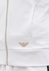 Λευκό μπουφάν εμπρός με χρυστό φερμουάρ και μικρό τρίγωνο με χρυσά καρφιά κοντά στην τσέπη.