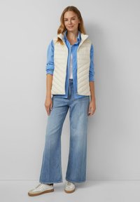 Mujer de pie con un chaleco acolchado crema sobre una camisa de botones azul, jeans de pierna ancha de color azul claro y zapatillas blancas, contra una pared gris.