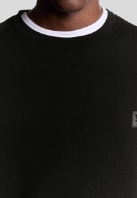 Felpa nera con collo rotondo, dotata di un colletto interno bianco, texture morbida e una piccola patch con logo sul fronte.