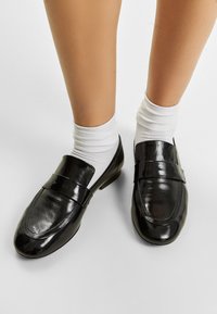 Mustat lakatut loafersit, joissa on sileä pinta, pyöristetty kärki ja koristeellinen hihna edessä, käytettynä valkoisten sukkien kanssa.