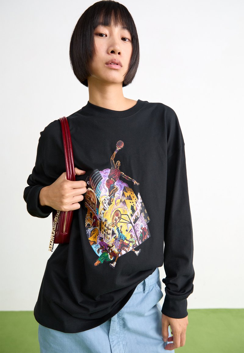 Camiseta de algodón negra de manga larga con un vibrante estampado gráfico que representa una escena de acción. Combinada con pantalones de mezclilla de color azul claro y un bolso rojo.