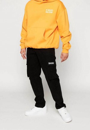 Felpa gialla con tasca frontale e testo stampato, abbinata a pantaloni cargo neri con tasche laterali e sneakers bianche.