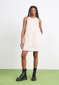 Vestido sin mangas de color beige con un corte relajado, que cuenta con bolsillos laterales y un pequeño logo rojo en el dobladillo. Combinado con botas negras de cordones.