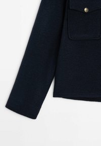 Manche d'un manteau en laine bleu marine avec poche à rabat et bouton doré décoratif sur fond blanc.