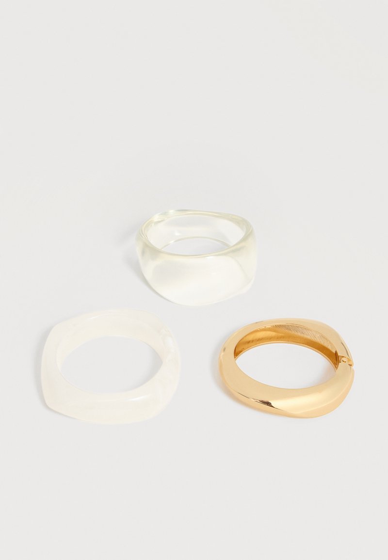 Trois anneaux incurvés en blanc, transparent et or disposés sur un fond clair uni.