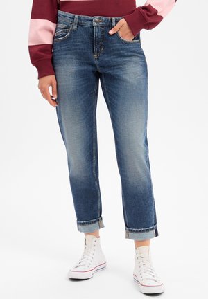 Person trägt hochgekrempelte blaue Jeans, weiße High-Top-Sneaker und ein bordeauxrotes und pinkes Langarmshirt, steht mit einer Hand in der Tasche.