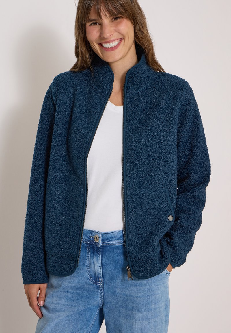 Jeune femme souriante portant une veste polaire bleu marine sur une chemise blanche avec un jean bleu clair, posant devant un fond clair uni.