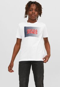 Balta kokvilnas t-kreklā ar tumši zilu un sarkanu grafisko dizainu, uz kura ir rakstīts "JACK & JONES", valkāta ar tumšām džinsiem. Vienkāršs apaļš kakla izgriezums un īsas piedurknes.