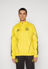 Mand iført en gul sportjakke med lynlås og sorte bukser med BVB 09 og Puma-logoer, der står mod en ensartet lys grå baggrund.