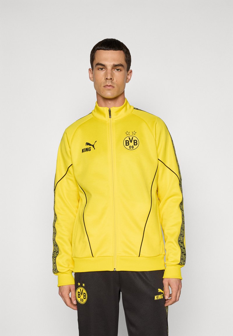 Mand iført en gul sportjakke med lynlås og sorte bukser med BVB 09 og Puma-logoer, der står mod en ensartet lys grå baggrund.