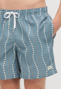 Swim trunks in light blue with wavy white lines and round polka dots. Features an elastic waistband with a drawstring and side pockets.