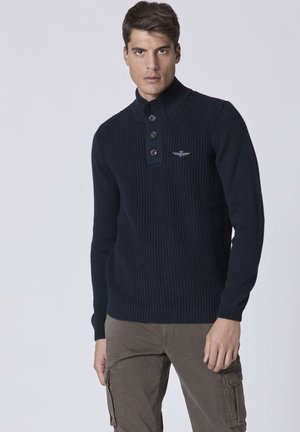 Maglione lavorato a maglia blu marino con colletto alto e pattina con tre bottoni, caratterizzato da un logo ricamato sul petto e polsini e orlo a coste.