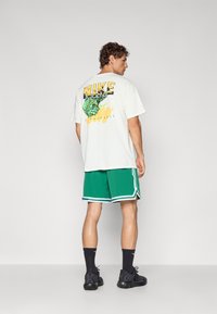 Hvid t-shirt med sort og gul grafisk tekst og grønt design på ryggen, parret med grønne shorts med hvide sidelister.