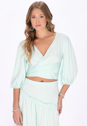 Tânără cu păr lung și ondulat purtând un top înfășurat verde mentă cu mâneci bufante, fustă asortată, cercei turcoaz și brățări la ambele încheieturi.