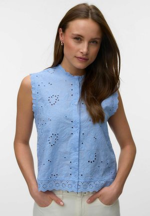 Femme aux longs cheveux bruns portant un chemisier sans manches bleu clair brodé d'œillets, debout les mains dans les poches d'un pantalon blanc.