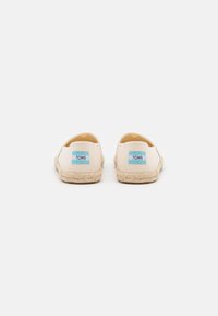Chaussures plates en toile beige clair avec une semelle en jute tressé. Présentent un patch logo TOMS bleu sur le talon et une finition lisse et texturée.