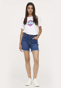 Shorts en denim avec un ourlet effiloché, dans un délavage bleu moyen. Assortis avec un t-shirt en coton blanc arborant un texte de logo coloré. Baskets blanches.