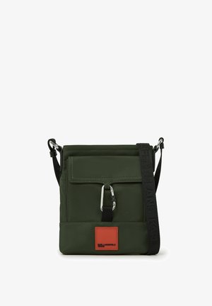 Zaļš neilona crossbody soma ar sudraba furnitūru, regulējamu siksnu, priekšējo atloku un brūnu ādas logo uzlīmi. Iezīmes ietver šuvju detaļu.