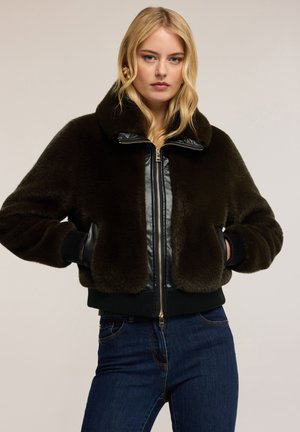 Kobieta z blond włosami, ubrana w ciemno-brązową kurtkę z sztucznego futra z czarnymi skórzanymi wykończeniami i niebieskie dżinsy, stoi z rękami w kieszeniach.