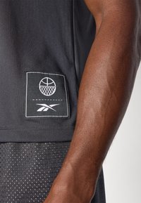 Schwarzes Basketball-Trikot mit einem Stoffaufnäher, der ein Basketballmotiv und das Reebok-Logo zeigt. Kurzärmlig, atmungsaktive Textur, leichtes Design.