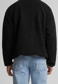 Czarna polarowa kurtka o fakturowanym materiale, w boxy kształcie i opuszczonych ramionach, zestawiona z jasnoniebieskimi dżinsami. Widoczne detale szwów na kieszeniach.