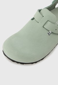 Sabot vert clair à enfiler avec une seule bride sur le dessus et une semelle noire texturée.