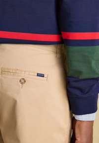 Pantalon beige avec poche arrière boutonnée étiquetée « Joules », associé à une manche de pull rayée marine, verte et rouge sur un poignet de chemise rayée.
