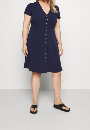 Mujer con un vestido azul marino hasta la rodilla con botones frontales, mangas cortas y sandalias tipo slide negras, de pie sobre un fondo blanco.