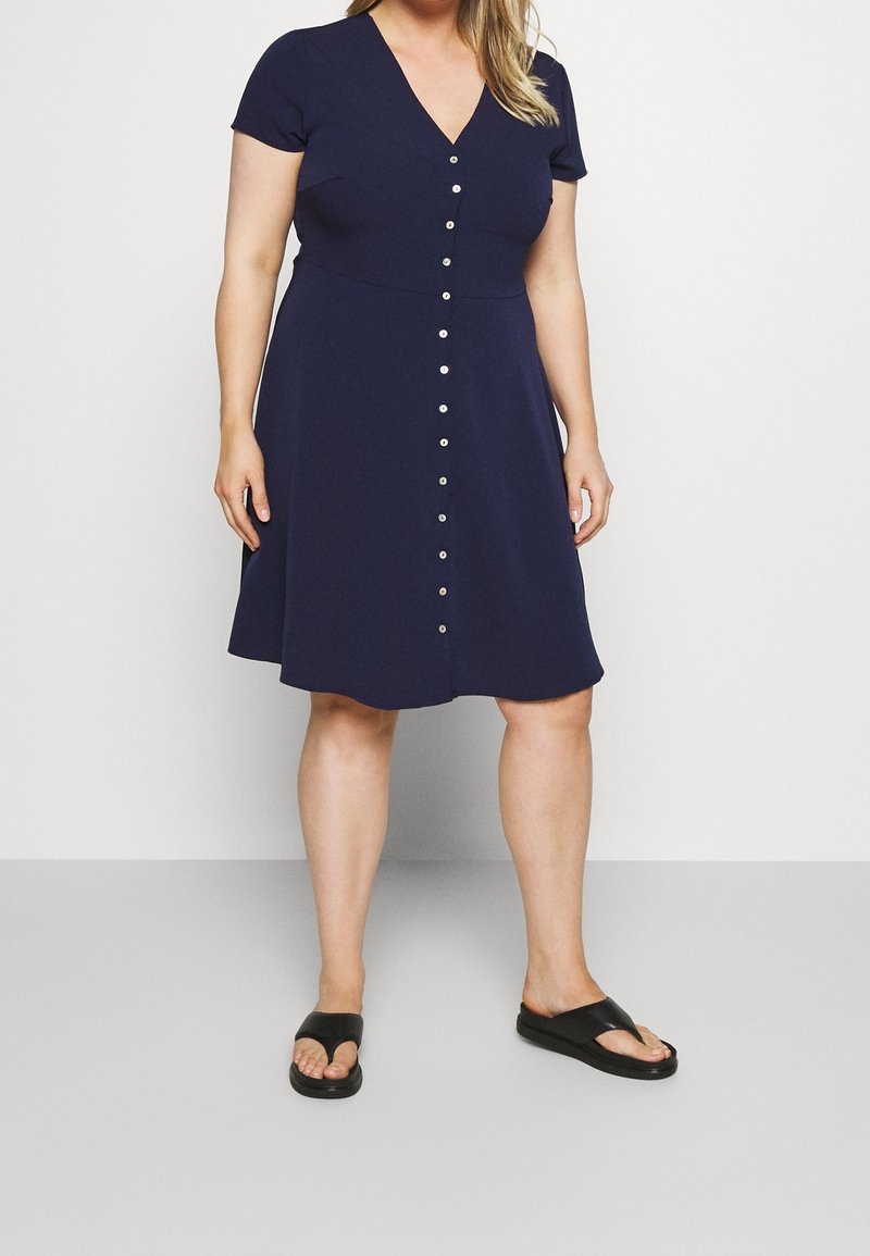 Femme portant une robe bleu marine longueur genou avec boutons devant, manches courtes et sandales slides noires, debout devant un fond blanc.
