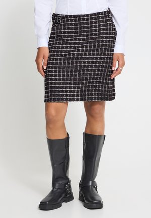 Jupe à carreaux noir et blanc portée avec des bottes noires montantes et une chemise blanche à manches longues, visible de la taille jusqu'aux pieds.