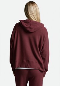 Eine blonde Person steht mit dem Rücken zur Kamera und trägt einen weinroten Hoodie und passende Hose, unter dem Hoodie ist ein weißes Shirt zu sehen.