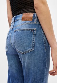 Jeans din denim albastru cu o textură decolorată, buzunare spate rotunjite și o etichetă din piele maro cu înscripția "TOMMY JEANS" embosată pe talie.