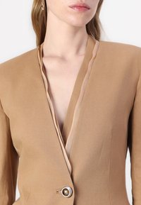 Bruine linnen blazer met een diepe V-hals, contrasterende beige afwerking en een enkele ronde knoopsluiting. Glad textuur en op maat gemaakte pasvorm.