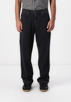 Melnas denim bikses ar vaļīgu piegriezumu, aprīkotas ar priekšējiem kabatām, vara kniedēm un vienkāršu dizainu. Valkātas ar melniem sporta apaviem.