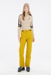 Beige Zip-Up Langarmtop mit schwarzen Streifen, kombiniert mit leuchtend gelben Skihosen. Schwarze Skischuhe vervollständigen das Wintersportoutfit.