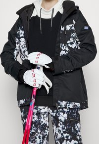 Person i svart skidjacka och byxor med blommönster, vita handskar, som håller i rosa skidstavar.