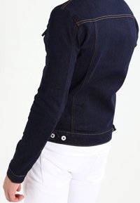 Person som bär en figursydd mörkblå denimjacka med knapppdetaljer, tillsammans med vita byxor, visad från sidan och baksidan.