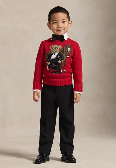 Jonge jongen draagt een rode trui met een design van een beer en een paard, zwarte broek, zwarte schoenen, wit overhemd en zwarte strik, staand tegen een neutrale achtergrond.