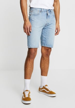 Shorts en denim bleu clair avec une coupe droite, ourlet effiloché, design à cinq poches et fermeture à bouton en métal. Portés avec des chaussettes blanches et des baskets marron.