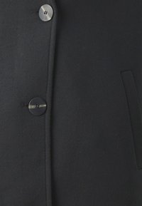 Vêtement d'extérieur noir en tissu texturé, avec une fermeture à boutons et des poches latérales. Le design inclut une silhouette épurée et ajustée.