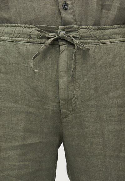 Κοντινό πλάνο ελιάς πράσινου λινών παντελονιού με μπροστινό κορδόνι δεμένο σε φιόγκο και ορατή ζώνη με κουμπιά.