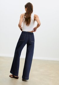 Široke temno modre denim hlače z visokim pasom, kontrastnim šivom in dvema zadnjima žepoma, v kombinaciji z oprijetim belim majico brez rokavov.