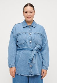 Light blue denim jacket with long sleeves, two front pockets, button closure, and a tied waist belt. Smooth texture, classic design.