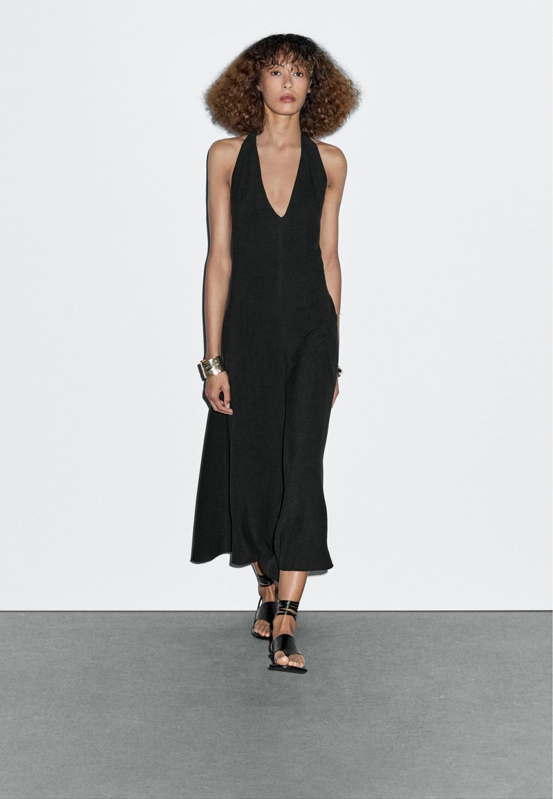 Jumpsuit nero con collo all'americana e profondo scollo a V, design elegante e gambe lunghe. Abbinato a sandali piatti neri e bracciale a manica metallico.