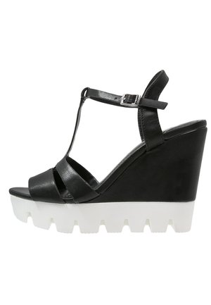 Schwarze Keilsandalette mit T-Riemen-Design, glattem Leder-Obermaterial und einer weißen Plattformsohle mit geriffelter Struktur. Verstellbarer Knöchelriemen.